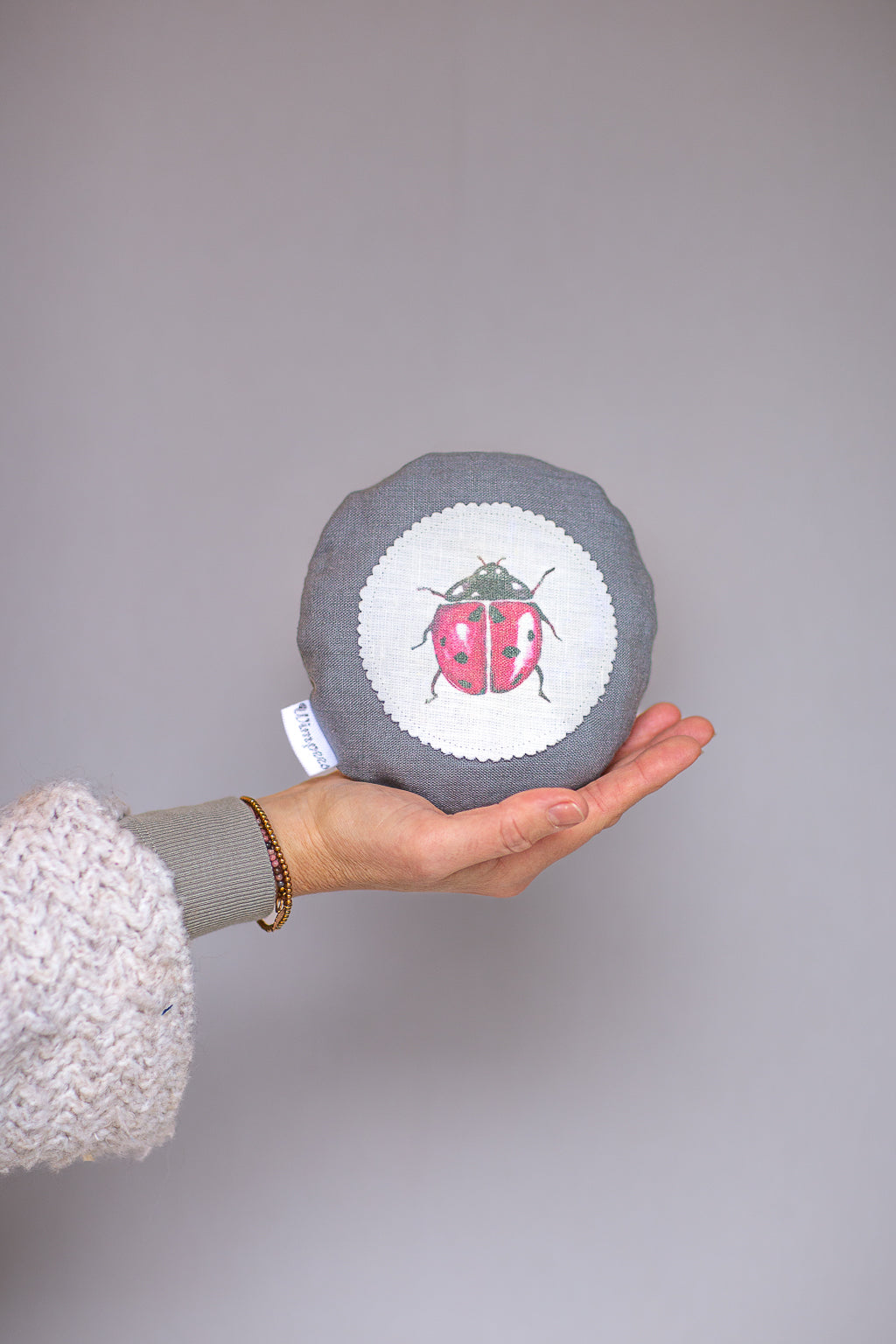 Rundes Dinkelkissen aus Leinen Farbe Grau mit Marienkäfer auf weißem Patch.