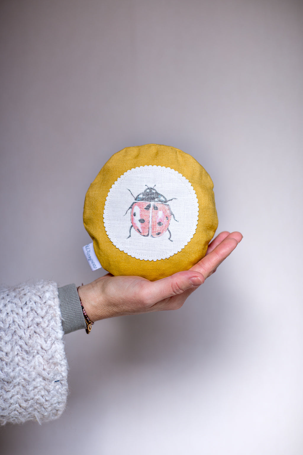 Rundes Dinkelkissen aus Leinen Farbe Senf mit Marienkäfer auf weißem Patch.